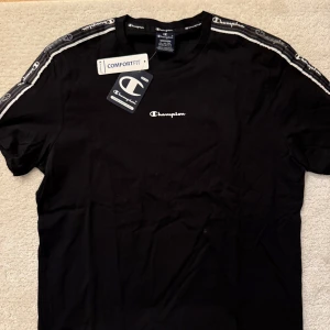 Svart t-shirt från Champion - Snygg svart t-shirt från Champion med logga på bröstet och längs ärmarna. Den har korta ärmar och en bekväm passform. Perfekt för en avslappnad stil. Storlek M. Helt ny.