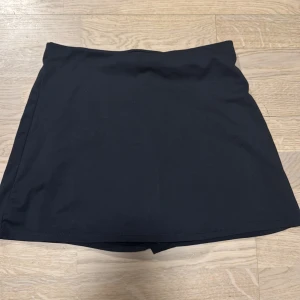 Svart kortkjol från Oysho - Aldrig använd Snygg svart kortkjol från Oysho, perfekt för en stilren look. Kjolen har en inbyggd shortsdesign för extra komfort och rörelsefrihet. Tillverkad i ett mjukt och stretchigt material.