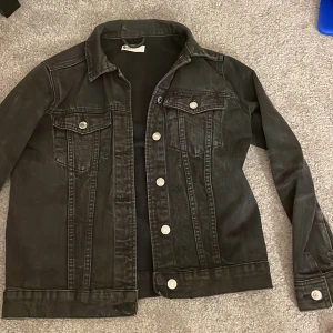 Svart jeansjacka - Snygg svart jeansjacka med klassisk design och silverfärgade knappar. Jackan har två bröstfickor med lock och knappar samt en rak passform. Perfekt för en cool och avslappnad stil.