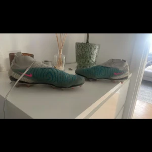 Nike fotbollsskor med blå och vit design - Snygga Nike fotbollsskor i en unik blå och vit färgkombination med rosa detaljer. Skorna har en modern design med snörning och är perfekta för fotbollsplanen.