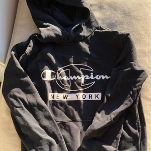 Svart hoodie från Champion - Säljer en svart hoodie från Champion med stor logga och texten 'New York' på framsidan. Den har en klassisk känguruficka och en bekväm huva. Perfekt för en avslappnad stil. Inget snöre kvar i huvan 