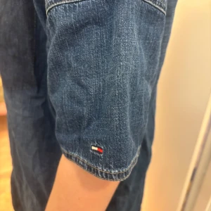 Jeansklänning från Hilfiger Denim - Snygg jeansklänning från Hilfiger Denim i en klassisk blå färg. Klänningen har korta ärmar och knappar framtill, vilket ger en avslappnad look. Perfekt för en casual stil med en touch av elegans.
