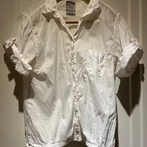 Vit skjorta med knappar - Klassisk vit skjorta med korta ärmar, knappar och bröstficka. Skjortan har långa ärmar som kan rullas upp och fästas med knapp. Perfekt för en stilren look.