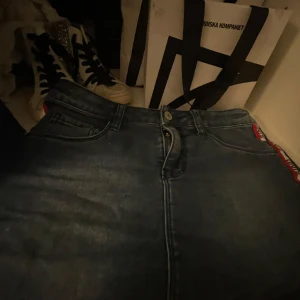 Blå kjol med Levis detaljer  - Snygg blå kjol  från Levis med en cool röd och vit rand längs sidan där Levis-loggan syns. Klassisk femficksdesign och knappgylf. Perfekta för en avslappnad stil med en twist.