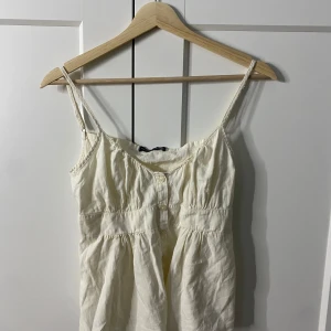 Vit linnetopp från Brandy Melville - Söt vit linnetopp från Brandy Melville med smala axelband och knappar framtill. Toppen har en lätt rynkad design och är perfekt för varma dagar.