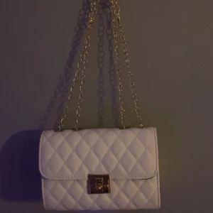 Vit quiltad axelväska med kedja - Snygg vit quiltad axelväska med guldfärgad kedja och låsdetalj. Perfekt för att ge en elegant touch till din outfit.