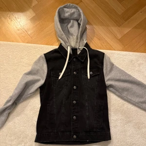 Svart jeansjacka med grå huva - Snygg svart jeansjacka med grå huva och ärmar i sweatshirtmaterial. Jackan har knappar framtill och två bröstfickor med lock. Perfekt för en avslappnad stil. Storlek S. Knappt använd och i väldigt bra skick.