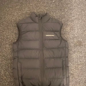 Svart dunväst från Peak Performance - Säljer en svart dunväst från Peak Performance med dragkedja framtill. Västen är perfekt för våren och sommaren 