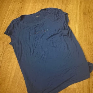 Blå t-shirt från Bebe - Snygg blå t-shirt från Bebe med glittrande stenar som bildar loggan på framsidan. Perfekt för en casual look med en touch av glamour. Väldigt stretchig 
