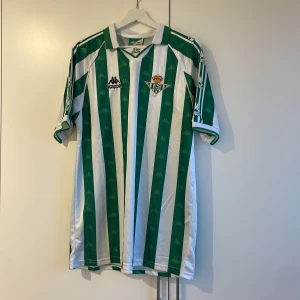 Grön och vit randig fotbollströja från Kappa - Säljer en grön och vit randig fotbollströja från Kappa med korta ärmar. Tröjan har klubbemblem på bröstet och Kappa-loggor längs ärmarna. Perfekt för fotbollsfans! Real betid hemma tröja 95/97