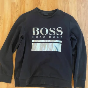 Svart tröja från Hugo Boss - Säljer en stilren svart tröja från Hugo Boss med stort tryck på framsidan. Tröjan har långa ärmar och rund halsringning, perfekt för en casual look.