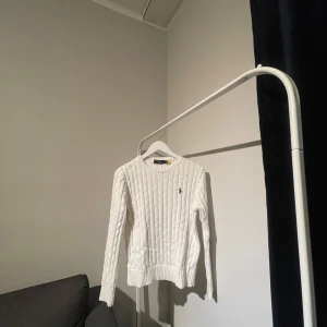 Vit stickad tröja från Ralph Lauren - Säljer en stilren vit stickad tröja från Ralph Lauren med kabelstickat mönster och broderad logga på bröstet. Tröjan har långa ärmar och rund halsringning. Perfekt för kyliga dagar! Använd 2 gånger
