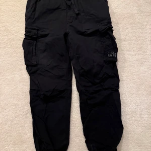 Svarta cargobyxor med fickor - Snygga svarta cargobyxor med flera praktiska fickor och snörning i midjan. Perfekta för en avslappnad stil och passar till många olika tillfällen. Baggy fit. Storlek M. Knappt använd.