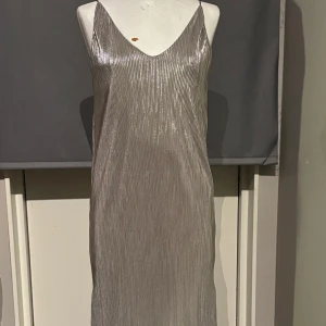 Glittrig silverklänning - Snygg glittrig silverklänning med v-ringad hals och smala axelband. Perfekt för en festlig kväll. Klänningen har en elegant och skimrande yta som fångar ljuset vackert.