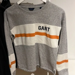 Grå tröja från Gant med ränder - Säljer en stilren grå tröja från Gant med vita och orangea ränder. Tröjan har en rund halsringning och långa ärmar. Nypris 999kr