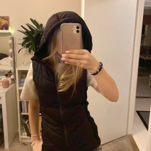 Svart dunväst med huva - Snygg svart dunväst med huva, perfekt för kyligare dagar. Västen har en dragkedja framtill och en figurnära passform som ger en stilren look. Ett mångsidigt plagg som funkar till många olika outfits.