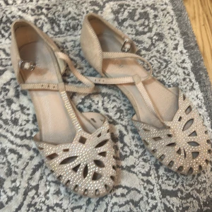 Beige sandaler med utskurna detaljer - Eleganta beige sandaler med utskurna mönster och glittrande detaljer. De har en smal rem över foten och en liten klack. Perfekta för barns festlighet.