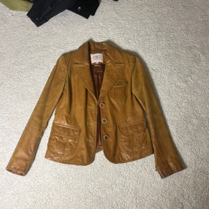 Brun skinnjacka från Blendshe - Snygg brun skinnjacka från Blendshe med klassisk krage och tre knappar framtill. Jackan har två fickor med lock och en bröstficka med knapp. Perfekt för en stilren look.