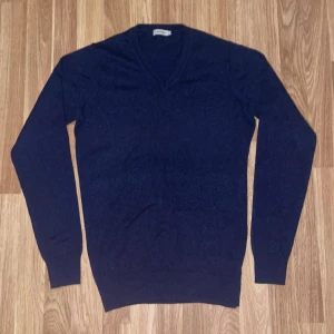 Mörkblå kashmirtröja från J.Lindeberg, 100% Kashmir - Säljer en elegant mörkblå kashmirtröja från J.Lindeberg. Tröjan har en v-ringad design och långa ärmar, perfekt för kyligare dagar. Den är tillverkad i 100% kashmir vilket ger en lyxig och mjuk känsla. Passar utmärkt för både vardag och mer uppklädda tillfällen.