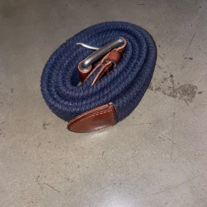 Flätat Ralph lauren bälte - Snyggt blått flätat bälte från Ralph Lauren med bruna läderdetaljer och en stilren silverfärgad spänne. Perfekt för att ge en marin touch till din outfit.