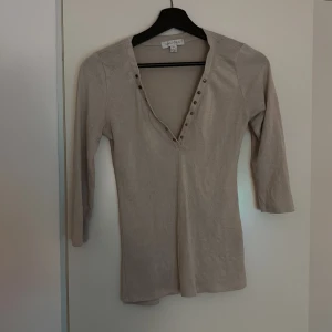 Beige topp från Magazine - Säljer en stilren beige topp från Magazine med trekvartsärmar och v-ringning dekorerad med knappar. Perfekt för en chic och avslappnad look.