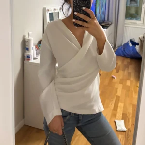 Vit omlottopp från Zara - Säljer en stilren vit omlottopp från Zara med långa ärmar och v-ringad design. Perfekt för en elegant look. Toppen har en draperad effekt framtill som ger en fin detalj.