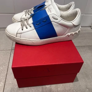 Valentino Garavani sneakers i vitt med blå rand strl 40 - Snygga Valentino Garavani sneakers i vitt med en bred blå rem över snörningen. Skorna har en unik design med nitar på hälen och en klassisk siluett. Kommer med låda och nya skosnören, Skorna är använda under vår och sommar. Skick 6-7/10