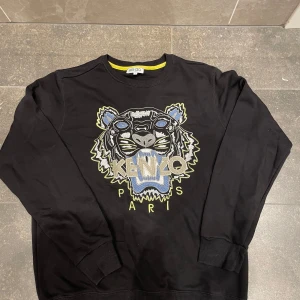 Svart sweatshirt från Kenzo med tigerbroderi - Snygg svart sweatshirt från Kenzo med ett stort broderat tigerhuvud och texten 'Kenzo Paris' på framsidan. Tröjan har långa ärmar och ribbade muddar vid ärmslut och nederkant. Perfekt för en stilren look.