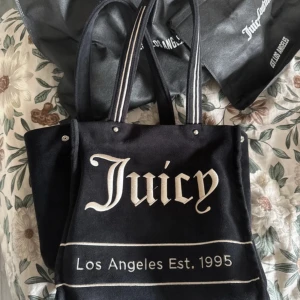 Svart shoppingväska från Juicy Couture - Snygg svart shoppingväska från Juicy Couture med texten 'Juicy Los Angeles Est. 1995' i vitt. Väskan har en praktisk dragkedja och kommer med en matchande liten vit plånbok med texten 'In Juicy We Trust'. Perfekt för att bära med sig allt man behöver.