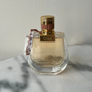 Parfym från Chloé - Elegant oanvänd parfym från Chloé med en gyllene kork och en ljusrosa vätska. Flaskan har en unik, rundad form och är dekorerad med ett brunt band runt halsen. Doftnoter: jasminblomma, fresia, lyxig vanilj, sandelträ och patchouli. 