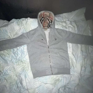 Grå huvtröja från Burberry - Snygg grå huvtröja från Burberry med klassiskt rutigt foder i huvan. Tröjan har dragkedja framtill och är tillverkad i bomullsmaterial. Perfekt för en stilren och bekväm look. Koftan är använd bara ett par gånger och säljs pga den är för stor på mig. 