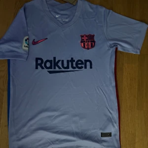 Ljusblå/lila FC Barcelona fotbollströja från Nike - Snygg ljusblå/lila FC Barcelona tröja från Nike med korta ärmar. Tröjan har klubbens emblem och sponsorlogga på framsidan samt La Liga-märke på ärmen. Perfekt för fans som vill visa sitt stöd.