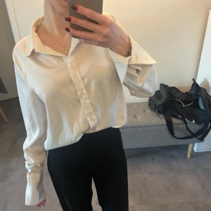 Vit satin skjorta med knappar - Säljer en klassisk vit skjorta i satin med knappar framtill. Skjortan har en normal passform och långa ärmar. Perfekt för en stilren look. Använd endast en gång från bik bok 