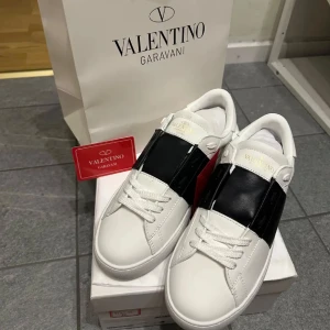 Valentino Garavani sneakers - Snygga Valentino Garavani sneakers i vitt med en bred svart rem över ovansidan. Skorna har snörning och små nitar längs sidorna för en extra touch. Perfekta för en stilren look. Skorna kommer utan låda osv