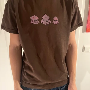 Brun t-shirt med tryck från Weekday - Säljer en brun t-shirt från Weekday med ett coolt tryck av tre rosa figurer på framsidan. T-shirten har en normal passform och är kortärmad, perfekt för en avslappnad stil.