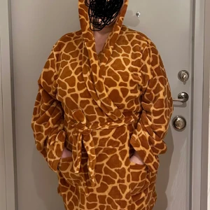 Giraffmönstrad badrock med huva - Mysig giraffmönstrad fleecejacka med huva och knytband i midjan. Jackan har långa ärmar och två praktiska fickor framtill. Perfekt för att hålla sig varm och bekväm hemma.
