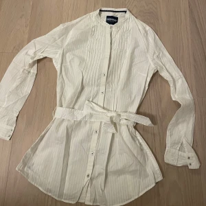 Vit långärmad skjorta från Sebago - Elegant vit långärmad skjorta från sebago med knappar och ett bälte i midjan. Skjortan har en diskret randig textur och en klassisk krage. Perfekt för en stilren look.