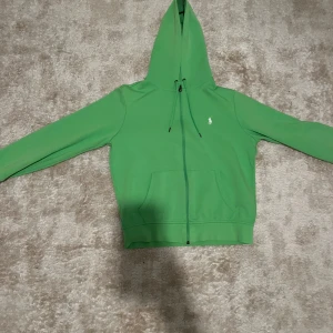 Grön hoodie med dragkedja - Snygg grön hoodie med dragkedja och huva. Den har en liten broderad logga på bröstet och praktiska fickor framtill. Perfekt för en avslappnad stil. Storleken är S, skicket är 8/10