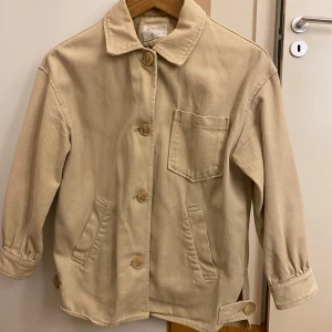 Beige jacka från Zara - Säljer en stilren beige jacka från Zara med knappar framtill och tre fickor. Jackan har en klassisk krage och justerbara remmar vid nederkanten. Perfekt för vår och höst!