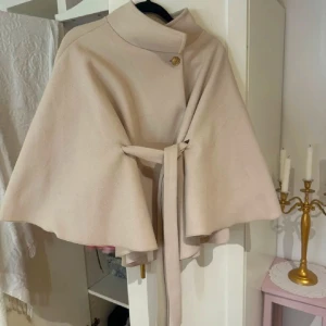 Beige kappa från Missay - Elegant beige kappa från Missay med knytband i midjan och en dekorativ knappdetalj vid halsen. Perfekt för en stilren look under hösten och våren 