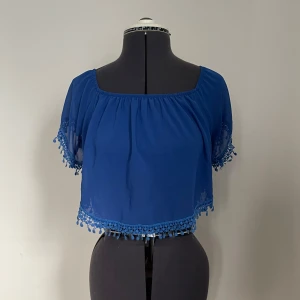 Blå croppad blus med spetsdetaljer - Blå croppad blus med spetsdetaljer och korta ärmar. Köpt utomlands så vet tyvärr ej vilket märke blusen är.