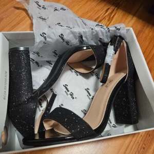 Svarta glittriga klackskor - Oanvända snygga svarta klackskor med glittrig yta och öppen tå. - storlek 37. - klackhöjd: 9,5cm. Skorna har en stabil klack och en elegant design.