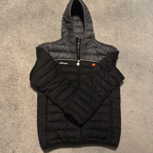 Svart och grå pufferjacka från Ellesse - Snygg pufferjacka från Ellesse i svart och grått med huva och dragkedja. Perfekt för kyligare dagar. Jackan har en broderad logotyp på bröstet och är långärmad.