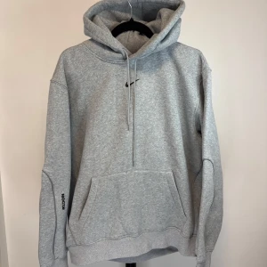 Nocta Grå hoodie från Nike - Säljer en klassisk grå hoodie från Nike med justerbar huva och känguruficka. Den har en liten Nike-logga på bröstet och är perfekt för en avslappnad stil. Passar bra till både träning och vardag.