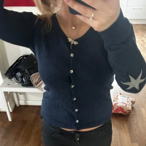 Mörkblå kofta med stjärndetalj - Säljer en mörkblå kofta med knappar framtill och en stjärndetalj på ärmen. Koftan har långa ärmar och en fin passform. Perfekt för att lägga till en touch av stil till din garderob.