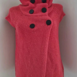 Rosa stickad kofta med stora knappar - Säljer en mysig rosa stickad kofta med stora svarta knappar. Koftan har en unik design med korta ärmar och en hög krage som kan vikas ner. Perfekt för att hålla sig varm och stilren.