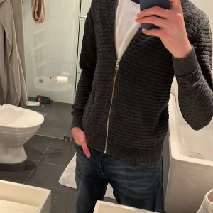 Grå kofta från GANT - Säljer en stilren grå kofta från GANT med dragkedja och ett diskret rutmönster. Koftan har långa ärmar och är perfekt för både vardag och mer avslappnade tillfällen. Den är tillverkad i ett mjukt material som ger en bekväm passform.