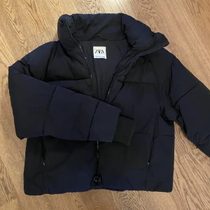 Svart pufferjacka från Zara - Säljer en svart pufferjacka från Zara med dragkedja och hög krage. Jackan har en stilren design och är perfekt för kyligare dagar. Den har långa ärmar och en bekväm passform. I bra skick!