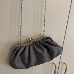 Grå kuvertväska med kedja - Elegant grå kuvertväska med en stilren kedja som axelrem. Väskan har en metallram och stängs med en klassisk bygelknäppning. Perfekt för en kväll ute eller en speciell tillställning.