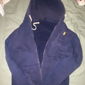 Mörkblå hoodie från Ralph Lauren - Säljer en snygg mörkblå hoodie från Ralph Lauren med dragkedja och huva. Den har en liten gul logga på bröstet och snören vid huvan. Perfekt för en avslappnad stil.